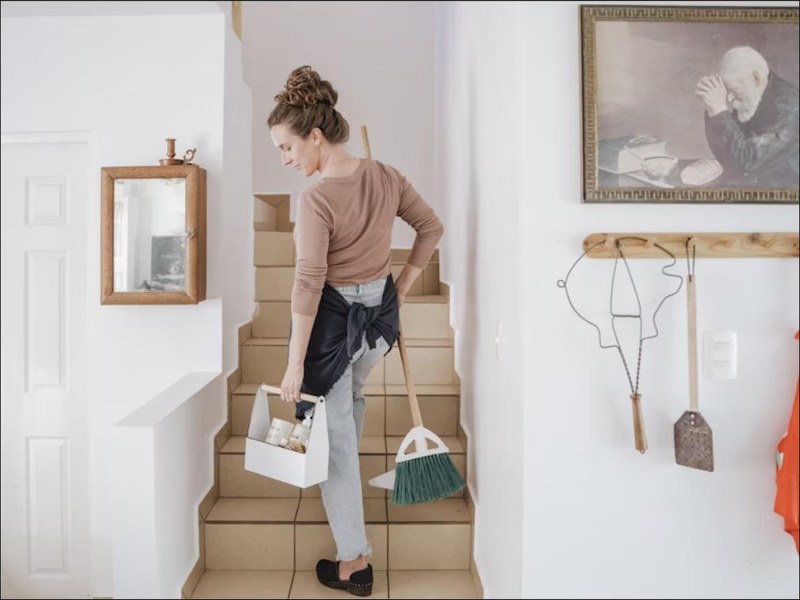 Cleaner carrying a caddy of supplies and a broom through a bright home